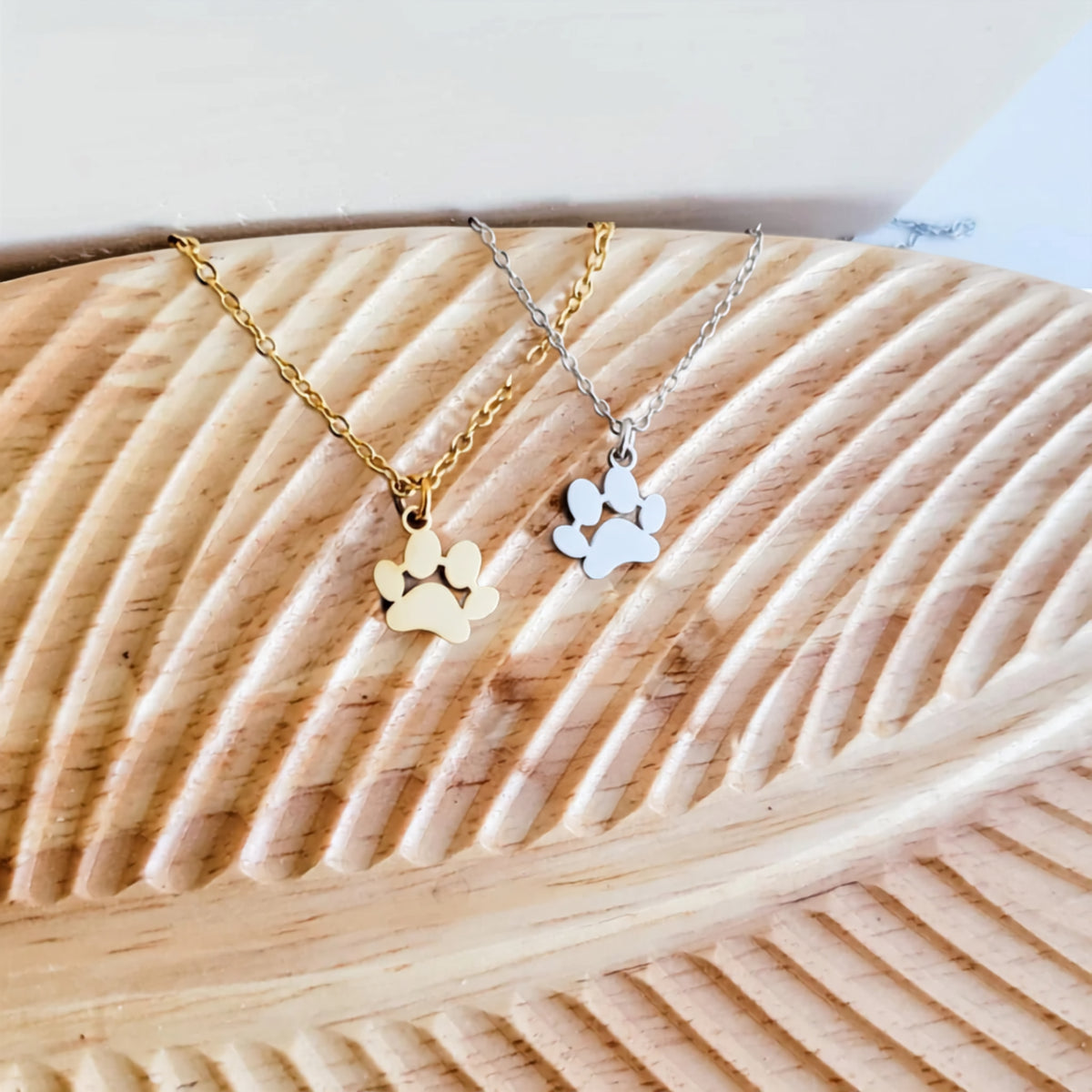 Paw print necklace in gold and silver stainless steel, draped over a wooden carved surface