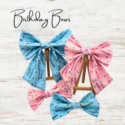 Pink and blue birthday-themed pet bows and bowties on a wooden stand with 'Birthday Bows' text above. The bows feature text saying "It's my barkday"