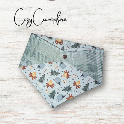 Reversible pet bandana with nature-themed pattern on a wooden surface, branded 'Cozy Campfire'. It has neutral colors for a chic look. 