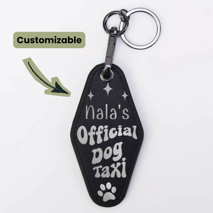 A black custom pet keychain shaped like a coffin, featuring the text 'Nala's Official Dog Taxi' in white lettering with a paw print at the bottom. It is shown with a silver split ring and clasp on a white background, with a 'Customizable' label and arrow graphic on the left.