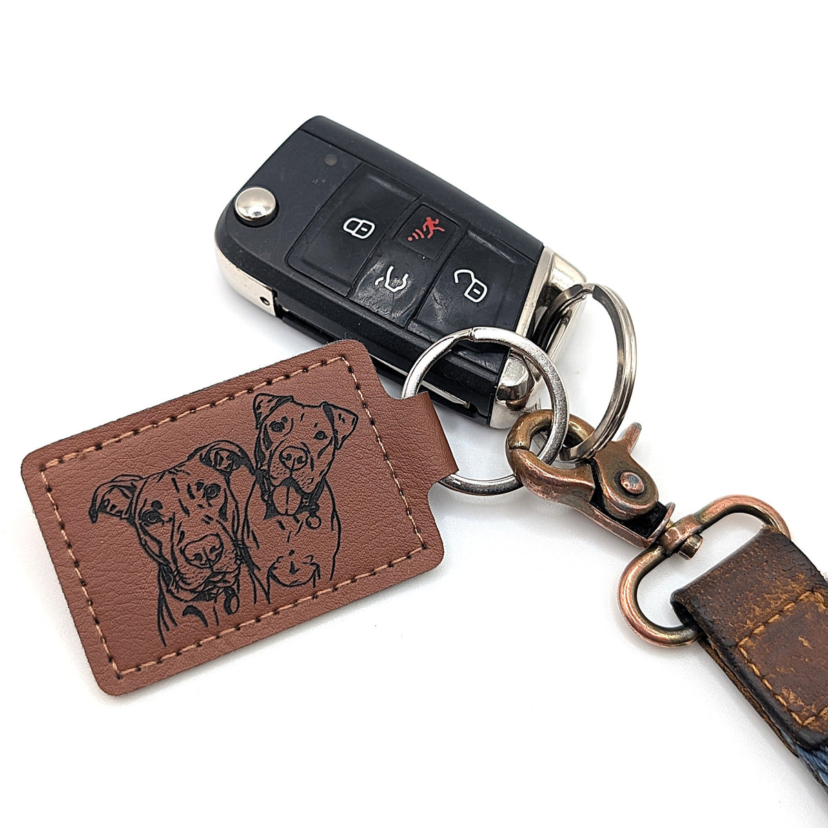 A brown leather keychain featuring an engraved portrait of two dogs on a rectangular tag, attached to a black car key fob and keyring hardware on a white background.