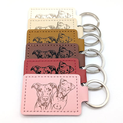Six dog keychains in different colors (cream, tan, brown, burgundy, red, and light pink) arranged in a vertical row, each with a stitched leather tag featuring a black line art illustration of two dogs and silver metal key rings.