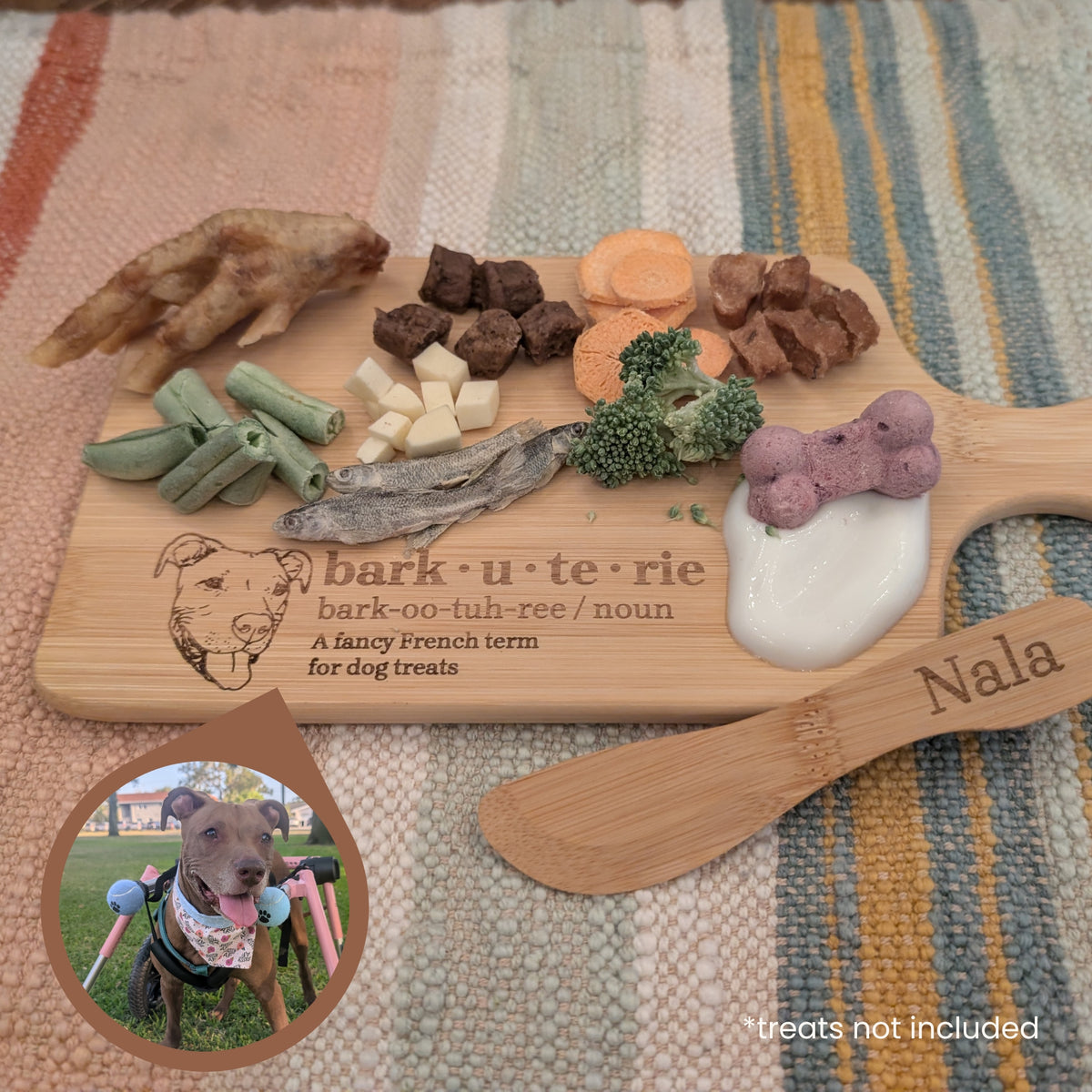 A wooden cutting board printed with the text 'bark-u-te-rie' and a dog illustration, styled as a flat-lay on a striped knit blanket. The board is loaded with various dog treats: brown cubes, carrot slices, green beans, dried fish, broccoli, a purple bone-shaped treat, and some yoghurt.