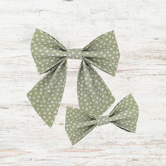 Two green geometrical patterned dog bows on a light wooden surface. One is bowtie style and the other is a girly sailor bow style