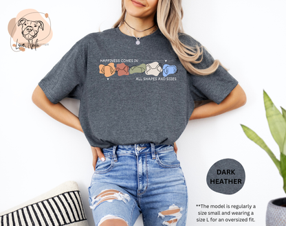 Person wearing a heather grey  t-shirt with a colorful graphic of dog ear outlines and text that says happiness comes in all shapes and sizes, standing against a white background.