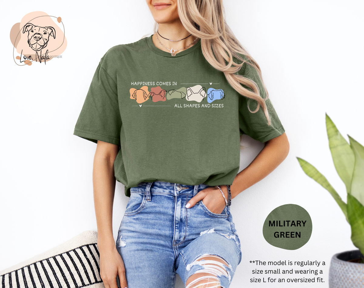 Person wearing a military green t-shirt with a colorful graphic of dog ear outlines and text that says happiness comes in all shapes and sizes, standing against a white background.