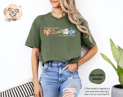 Person wearing a military green t-shirt with a colorful graphic of dog ear outlines and text that says happiness comes in all shapes and sizes, standing against a white background.