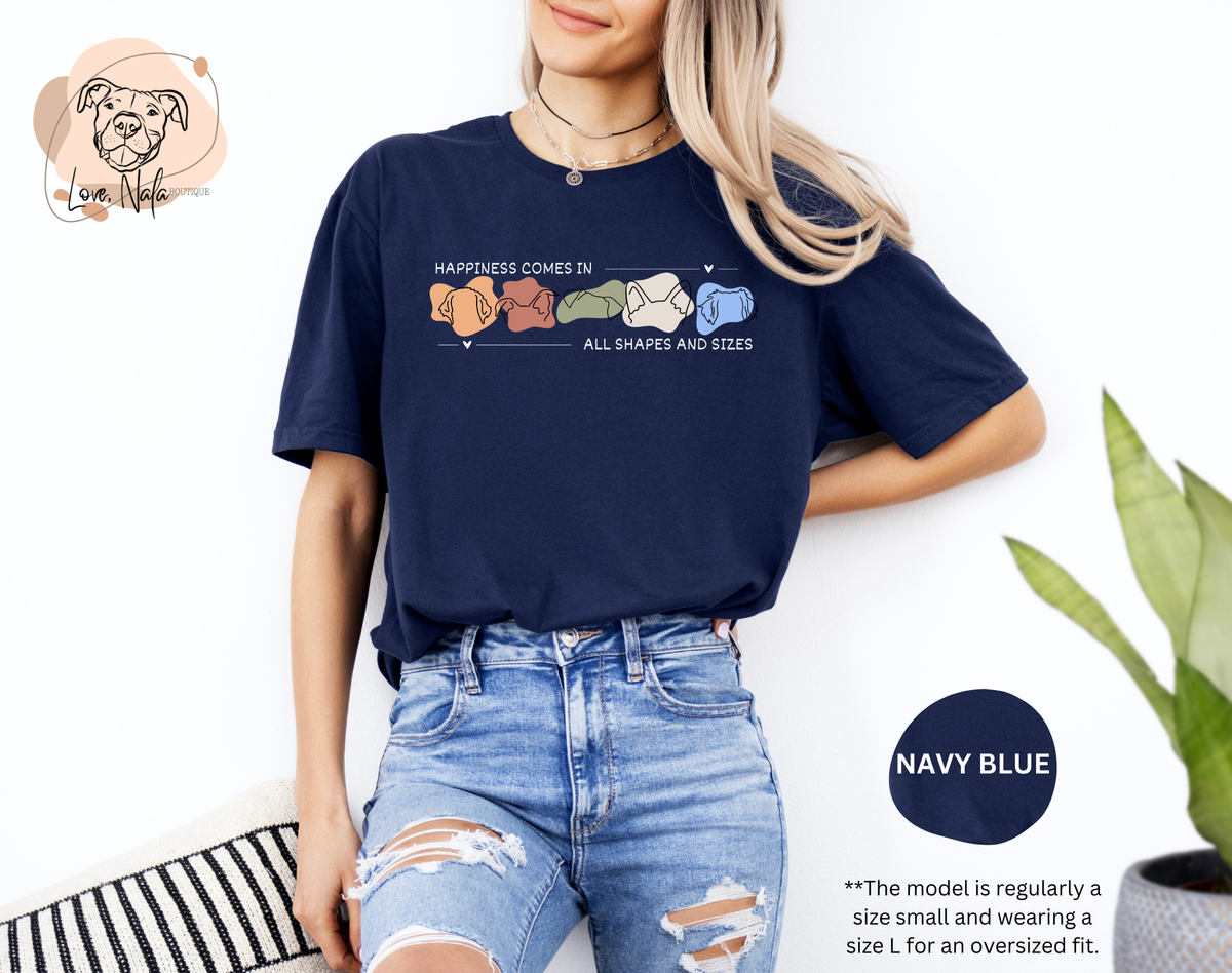 Person wearing a navy blue t-shirt with a colorful graphic of dog ear outlines and text that says happiness comes in all shapes and sizes, standing against a white background.
