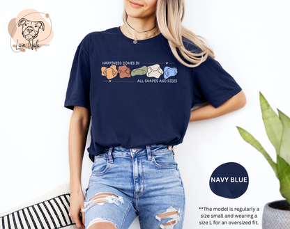 Person wearing a navy blue t-shirt with a colorful graphic of dog ear outlines and text that says happiness comes in all shapes and sizes, standing against a white background.
