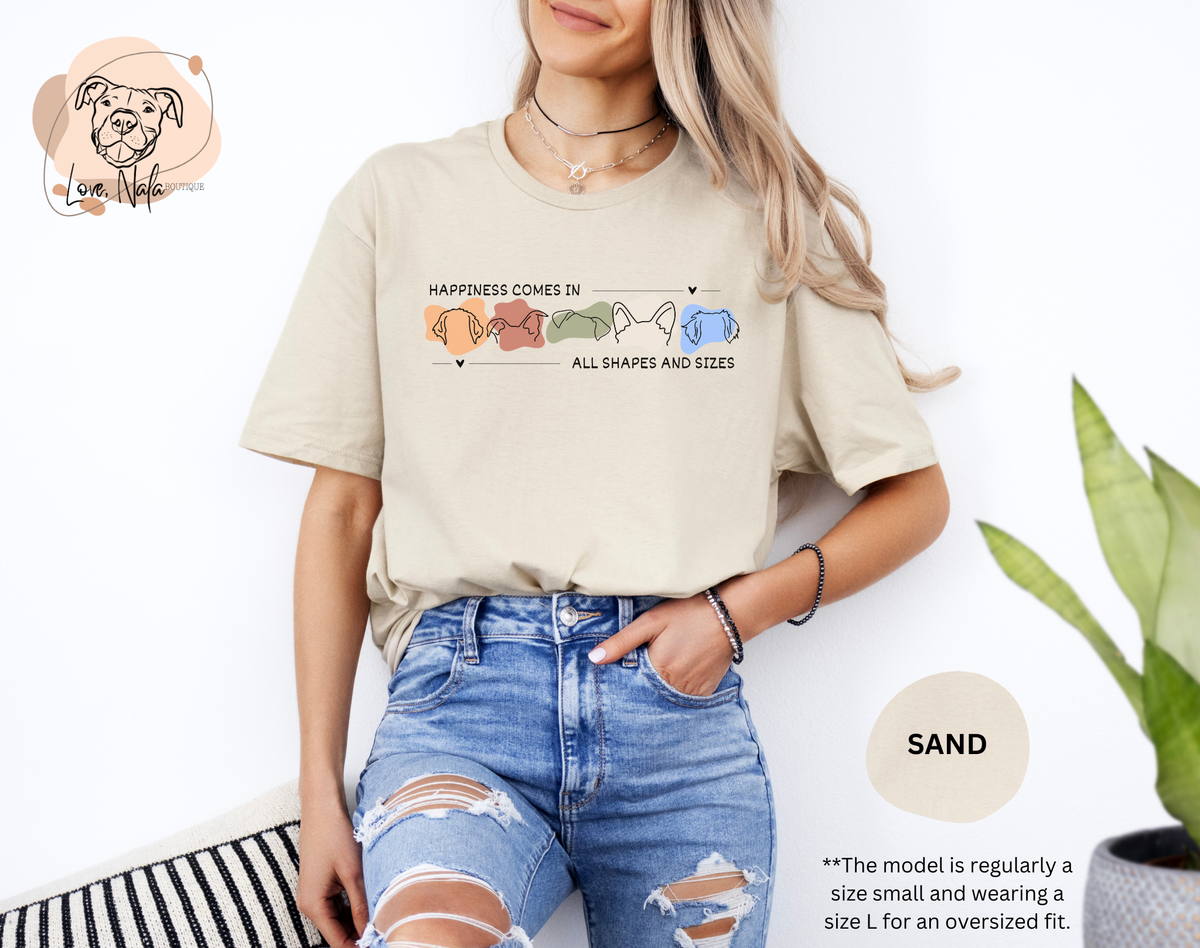 Person wearing a sand color t-shirt with a colorful graphic of dog ear outlines and text that says happiness comes in all shapes and sizes, standing against a white background.