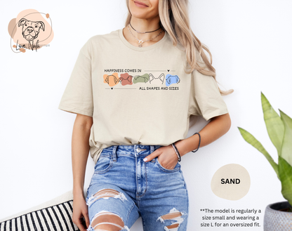 Person wearing a sand color t-shirt with a colorful graphic of dog ear outlines and text that says happiness comes in all shapes and sizes, standing against a white background.
