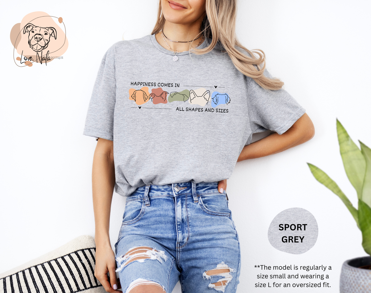 Person wearing a sport grey t-shirt with a colorful graphic of dog ear outlines and text that says happiness comes in all shapes and sizes, standing against a white background.