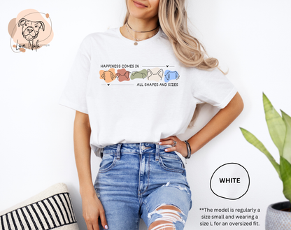 Person wearing a white t-shirt with a colorful graphic of dog ear outlines and text that says happiness comes in all shapes and sizes, standing against a white background.