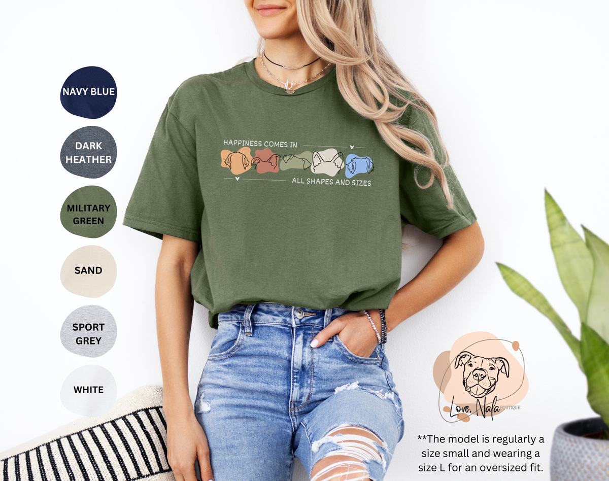 Person wearing an olive green t-shirt with a colorful graphic of dog ear outlines and text that says happiness comes in all shapes and sizes, standing against a white background.