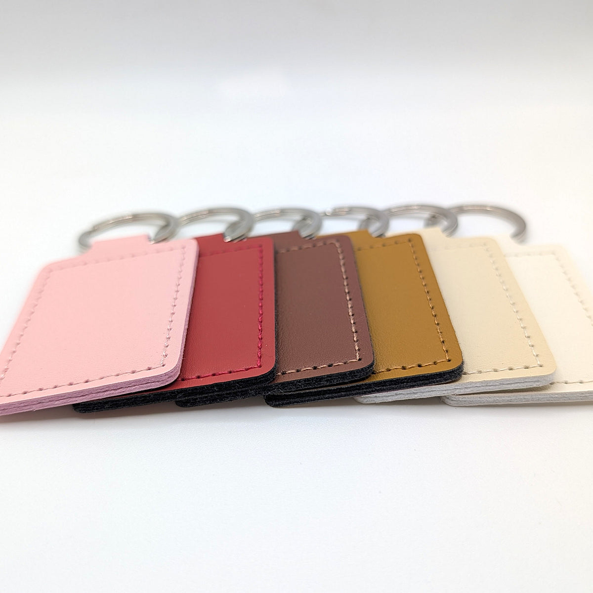 Six rectangular leather keychain tags in pink, red, brown, tan, white, and beige, arranged in a row with silver metal key rings on a white background.