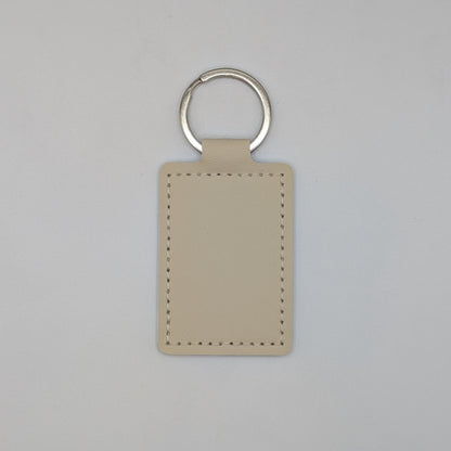 A minimalist beige leather keychain tag with a silver split ring, shown against a neutral light gray background.