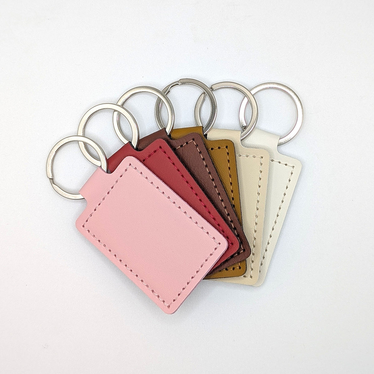 Five rectangular leather keychains in pink, burgundy, brown, tan, and cream, each attached to a silver metal key ring, arranged in a fan shape on a white background.