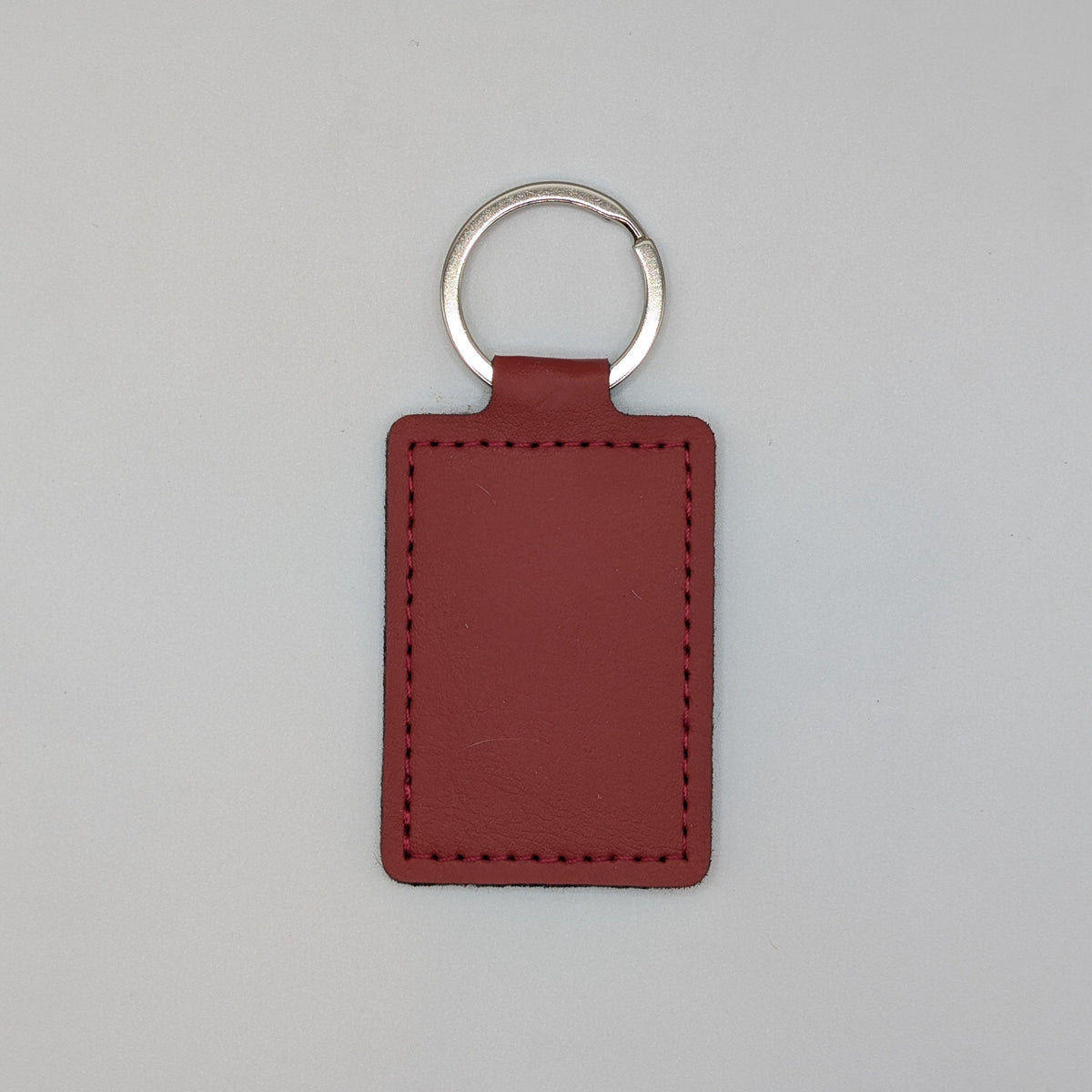 A rectangular leather keychain tag in a reddish-brown color with a silver metal split ring, shown against a plain light gray background.