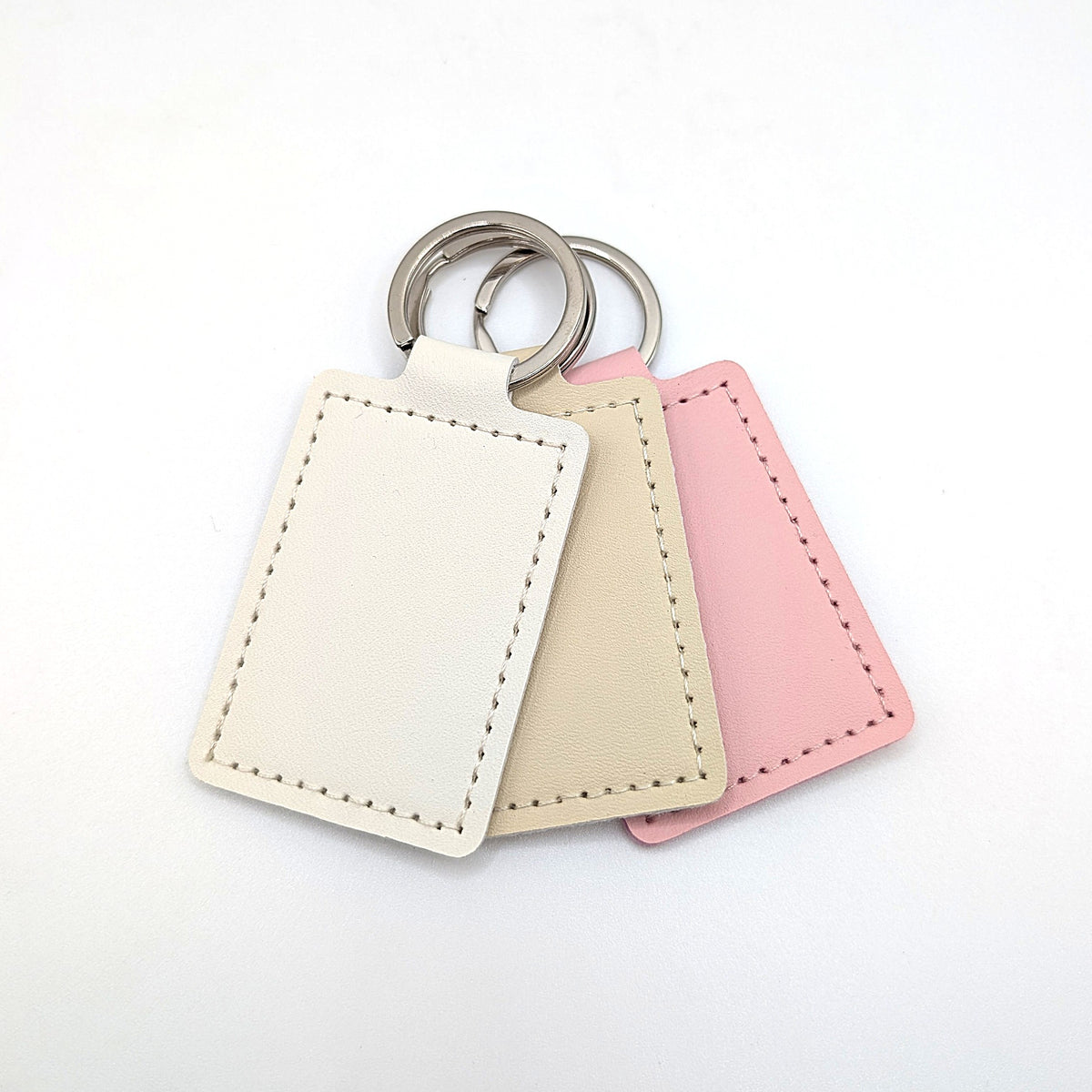 Three rectangular leather keychains in cream, beige, and pink, arranged in a fan shape with silver metal key rings on a white background.