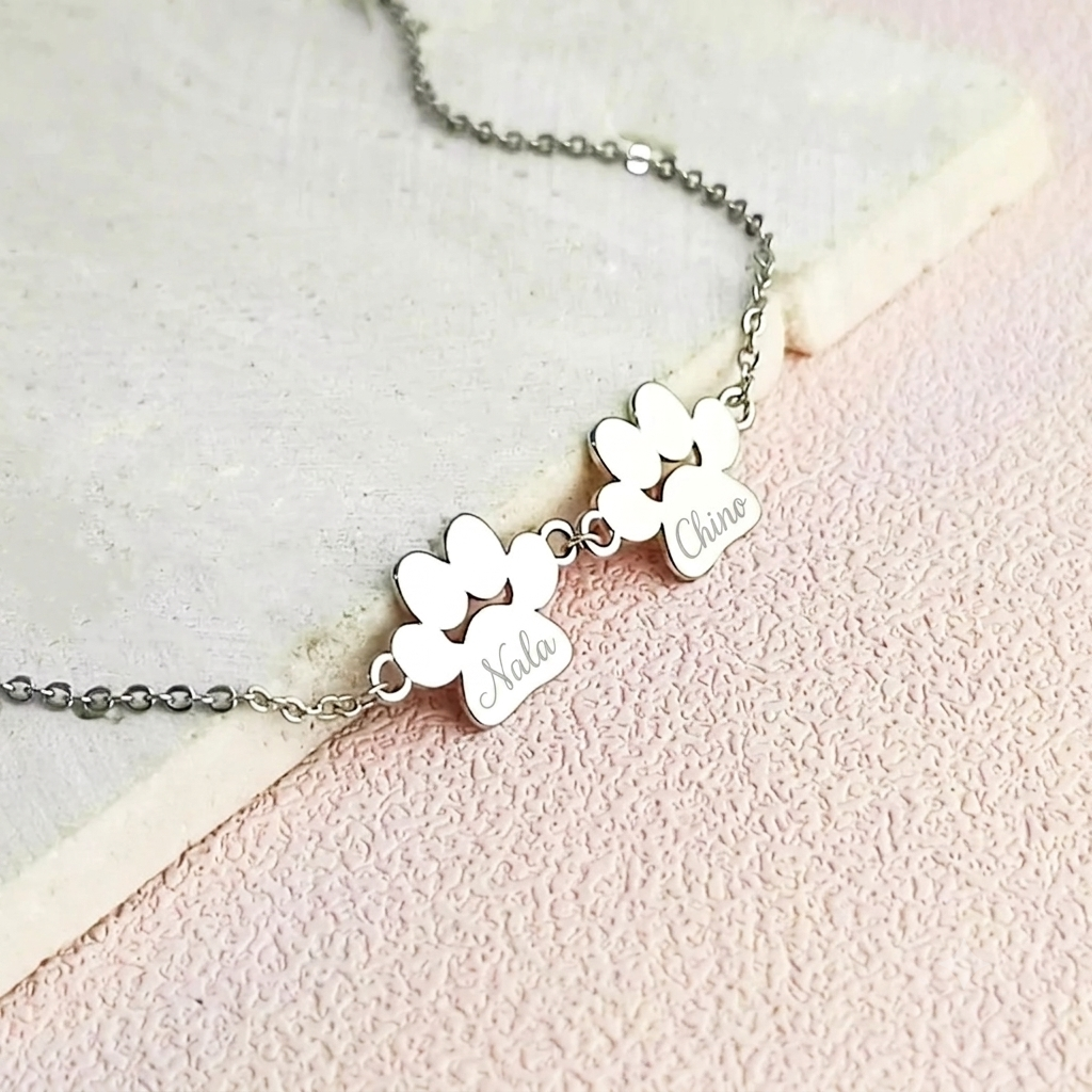Silver bracelet with two engraved paw prints shaped pendants that have 'Nala' and 'Chino' engraved on them. The jewelry is laying on a light pink background