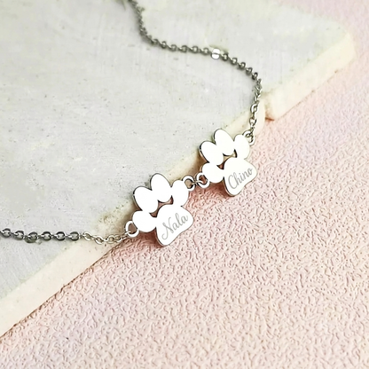 Silver bracelet with two engraved paw prints shaped pendants that have 'Nala' and 'Chino' engraved on them. The jewelry is laying on a light pink background