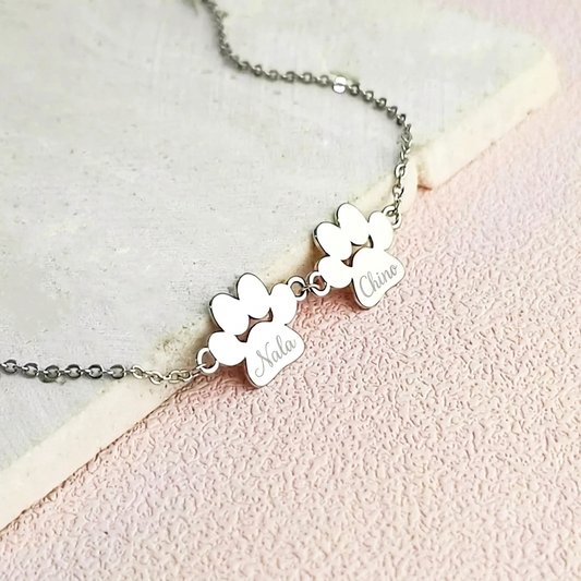 Silver bracelet with two engraved paw prints shaped pendants that have 'Nala' and 'Chino' engraved on them. The jewelry is laying on a light pink background