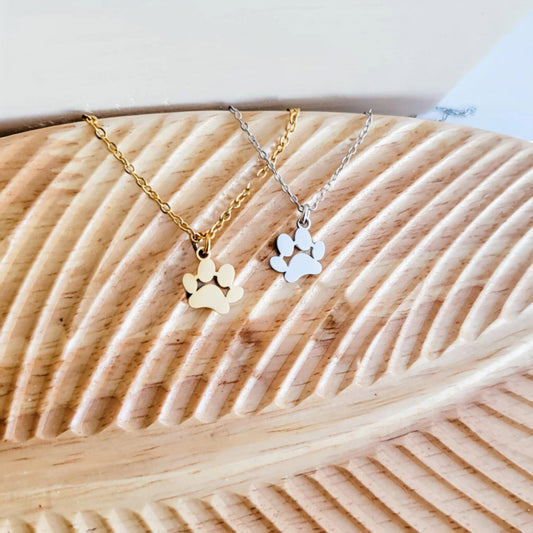 Paw print necklace in gold and silver stainless steel, draped over a wooden carved surface