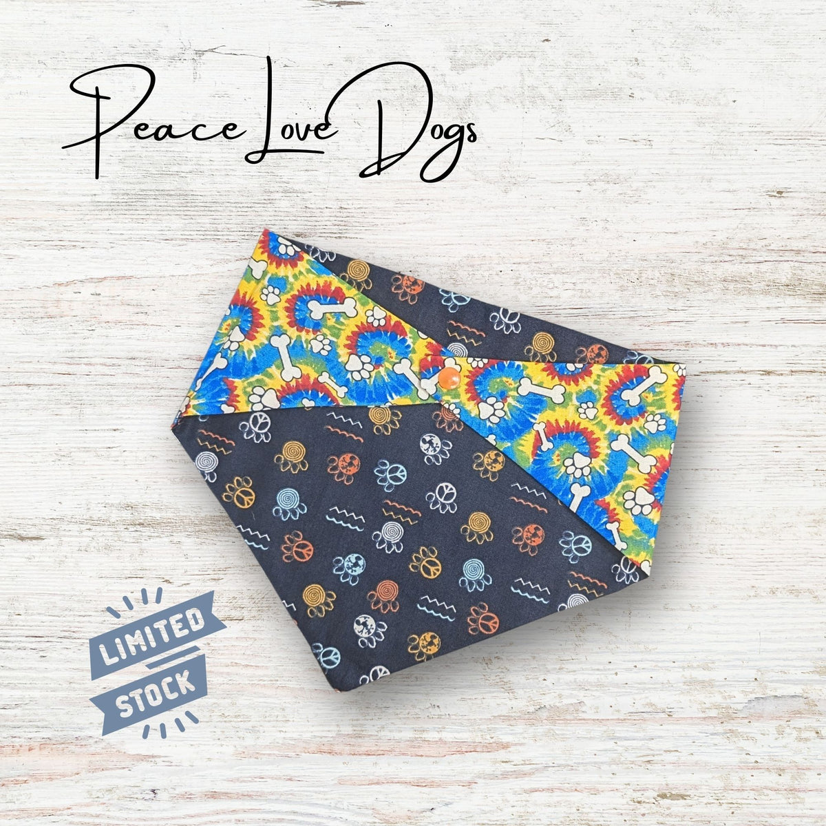 Two sided patterned bandana with 'Peace Love Dogs' branding on a light wooden surface. The fabric is a tie dye with paw prints and bones, the other size is paw prints that have peace signs inside