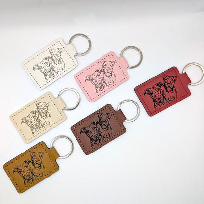 Six leather keychains in different colors (beige, pink, brown, and red) arranged on a white background. Each keychain features an engraved illustration of two dogs and is attached to a silver metal split ring.