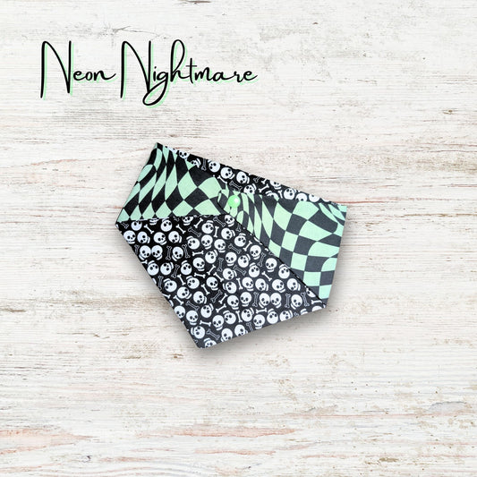 Two patterned pet bandanas on a light wooden surface with 'Neon Nightmare' text. The bandana has skulls and bones on one side with a trippy green checkered print on the reverse side