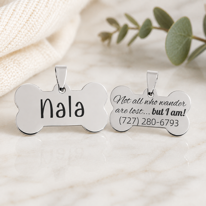 Two silver bone-shaped pet ID tags on a light marble surface with a cream knit fabric and green leaves in the background. One tag is engraved with the name 'Nala' and the other with the phrase 'Not all who wander are lost.