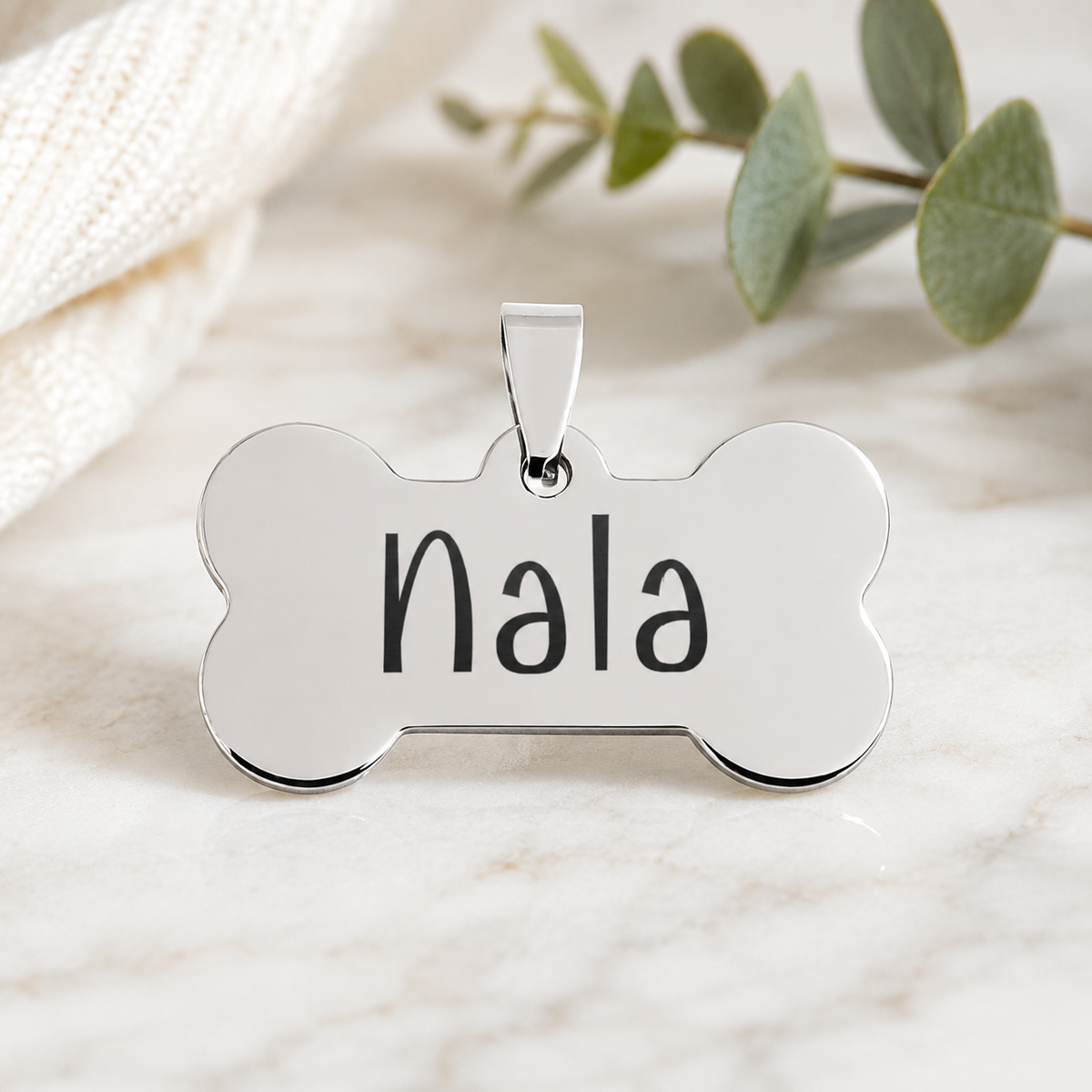 A polished silver-tone bone-shaped pet ID tag engraved with the name 'Nala' in black lettering, resting on a light marble surface with a soft cream fabric and green eucalyptus leaves in the background.