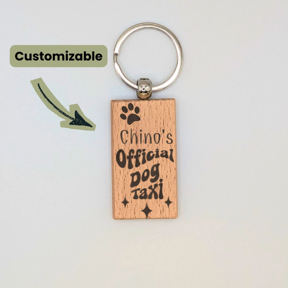 A rectangular wooden keychain with a silver metal key ring, featuring the engraved text 'Chino's Official Dog Taxi' and a paw print. An arrow graphic points to the keychain with a 'Customizable' label on a plain light background.