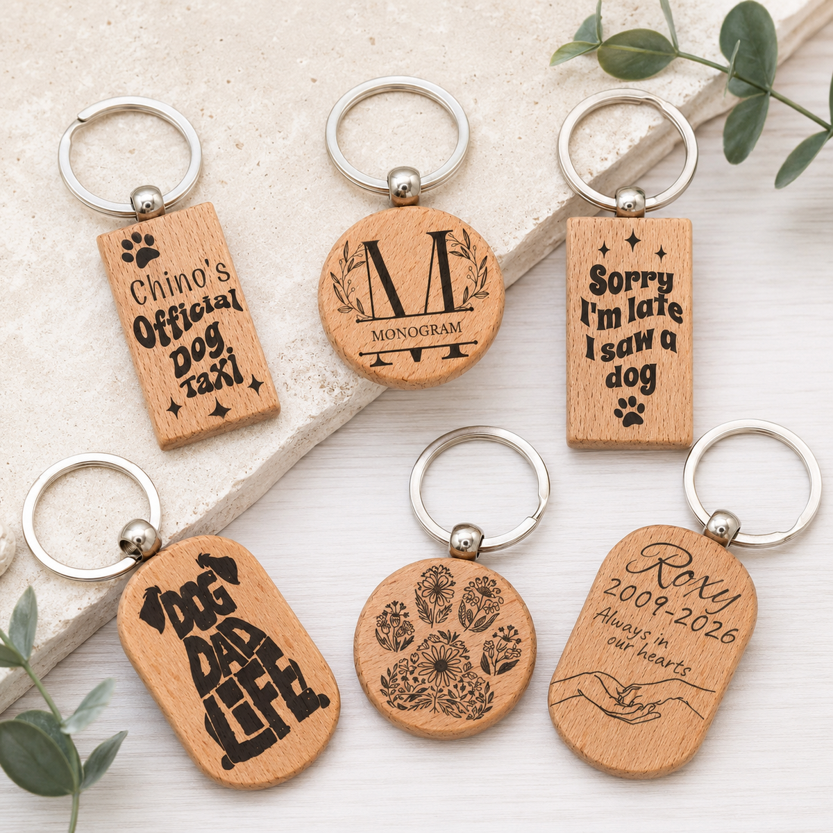 Six personalized wooden keychains with silver metal key rings, arranged on a light stone slab and white wood surface with eucalyptus leaves. The keychains feature engraved designs including a dog silhouette, paw prints, a monogram 'M', and text such as 'Chino's Official Dog Taxi', 'Sorry I'm late I saw a dog', and 'Rory 2009-2026 Always in our hearts'.